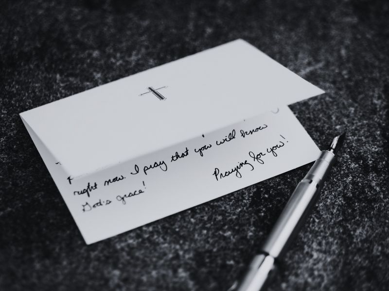 A prayer note card with a Christian cross on top of the card, lies partly open showing a personal note written inside the card; a fountain pen is beside the card. Photo by Aaron Burden on Unsplash aaron-burden-gV0l1uYr98M-unsplash. Photo was edited from colour to black and white in Canva by Siteri R.