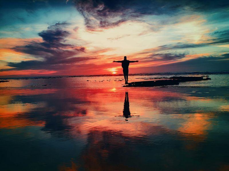 A person stands at a beach facing a colourful sunset and throws their arms wide open praising God.