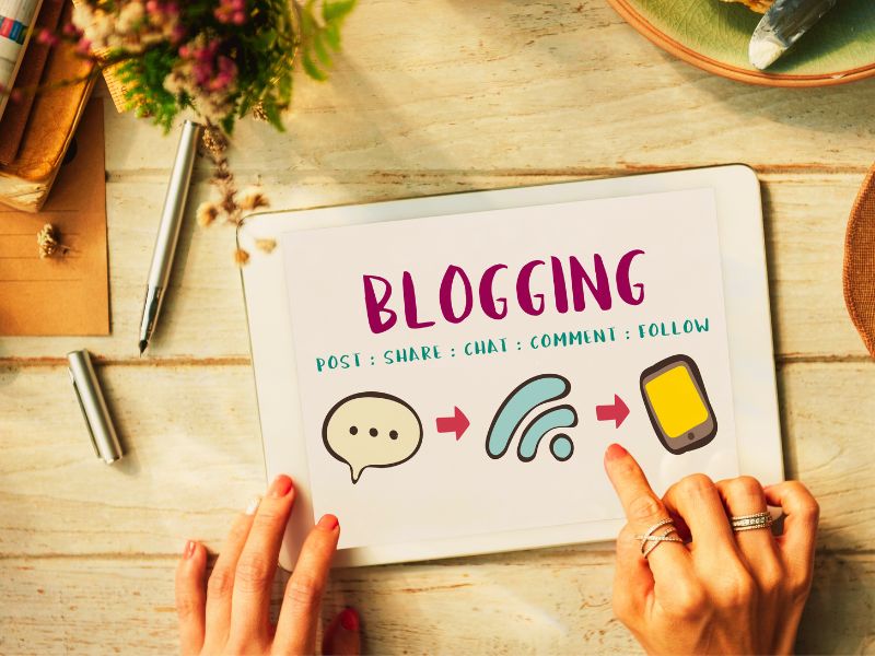 A white digital smart tablet is on a wooden table and shows the word 'BLOGGING' on the screen. Photo by PXHere.